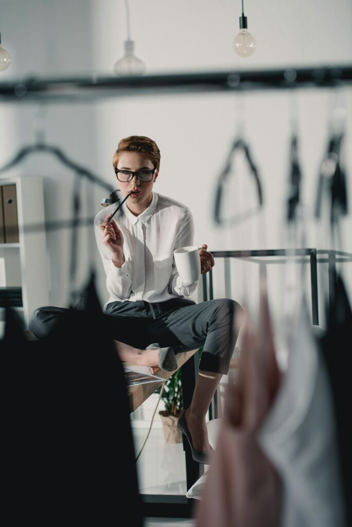 stylish fashion designer with cup of coffee sitting on desk and looking at dresses